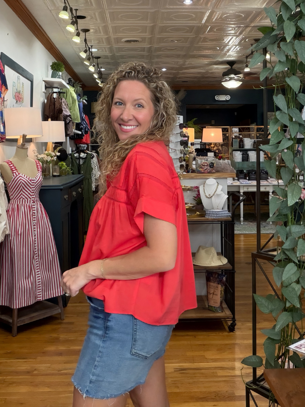 A woman with curly hair wearing a bright coral-red ruffled short-sleeve blouse and a denim skirt, posing inside a boutique shop.