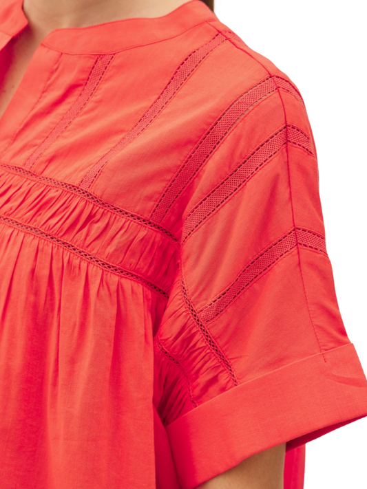 Close-up of a bright coral-red short-sleeve top with gathered smocked detailing and decorative contrast stitching on the sleeve and chest, shown on a model against a white background.