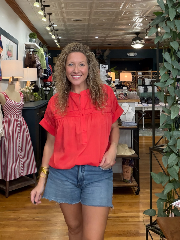 A smiling woman with curly hair stands in a boutique wearing a bright coral-red short-sleeve blouse with a gathered neckline and blue denim shorts. The shop has wood floors, a decorative ceiling, clothing racks, and potted plants.
