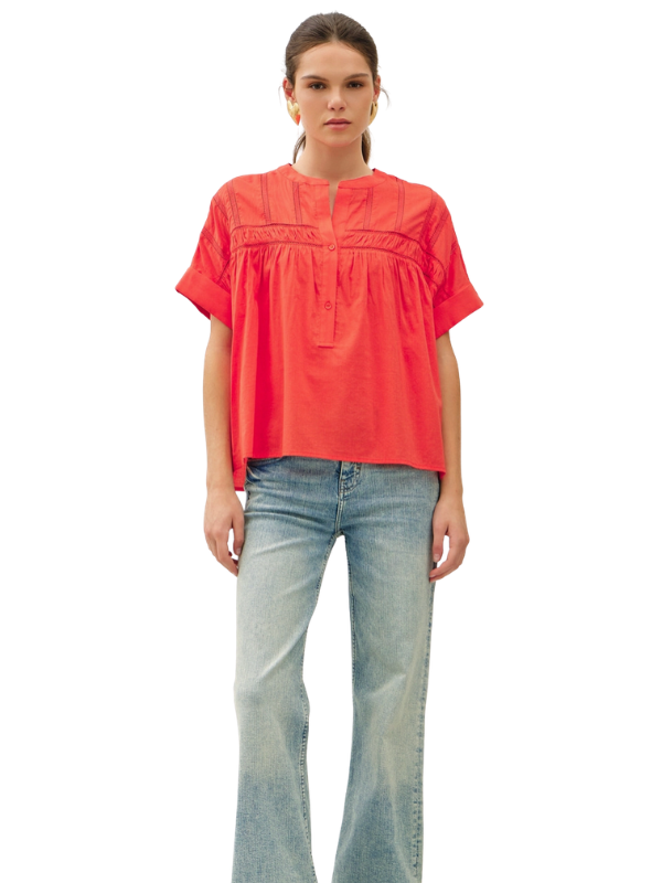 A woman modeling a short-sleeve, vibrant coral-red blouse with gathered detailing at the chest, paired with light-wash blue jeans, against a plain white background.