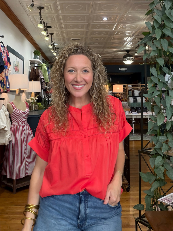 A smiling woman with curly blonde hair wearing a bright coral-red short-sleeve blouse and blue jeans, standing inside a boutique store with clothing racks, lamps, and a potted plant visible in the background.