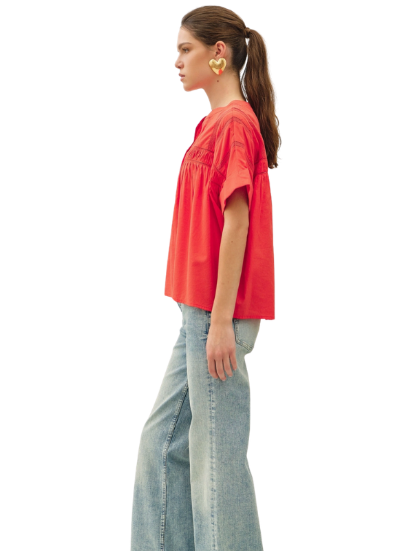A woman in profile wearing a bright coral-red short-sleeve top and light-wash wide-leg jeans against a plain white background.