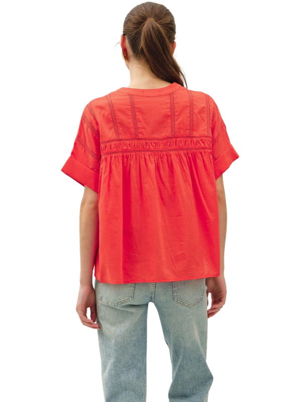A woman viewed from the back wearing a bright coral-red short-sleeve blouse with gathered detailing across the upper back and light-wash jeans, against a plain white background.