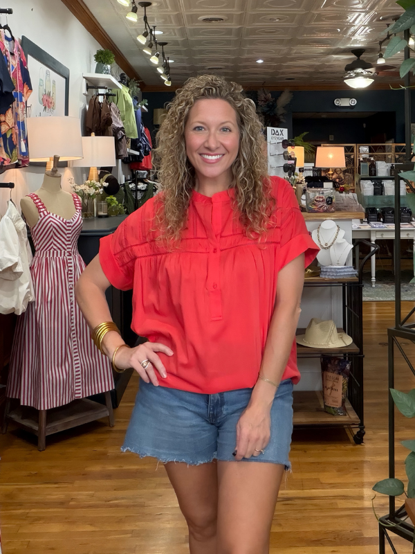 A smiling woman with curly hair stands inside a clothing boutique wearing a bright coral-red short-sleeve blouse and denim shorts. Mannequins and store fixtures are visible in the background.