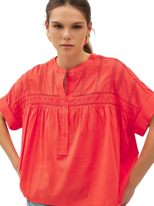A woman wearing a bright coral-red short-sleeve blouse with vertical gathered detailing and a split neckline, shown against a plain white background.