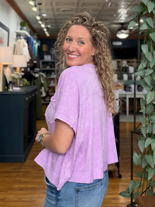 A smiling woman with curly blonde hair wearing a light purple, acid-washed short-sleeve t-shirt with a chest pocket, paired with blue jeans, standing inside a retail store.