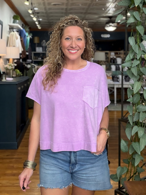 A woman with curly blonde hair stands in a retail store wearing a light purple short-sleeve crewneck top with a chest pocket and blue denim shorts.