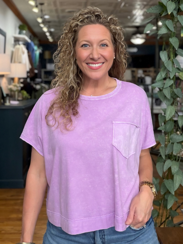 A smiling woman with curly blonde hair wearing a light purple, acid-washed short-sleeve t-shirt with a chest pocket, paired with blue jeans, standing inside a retail store.