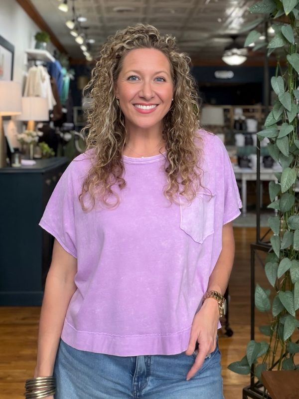 A smiling woman with curly blonde hair wearing a lavender short-sleeve crewneck sweatshirt with a chest pocket, paired with blue jeans, standing inside a retail store.