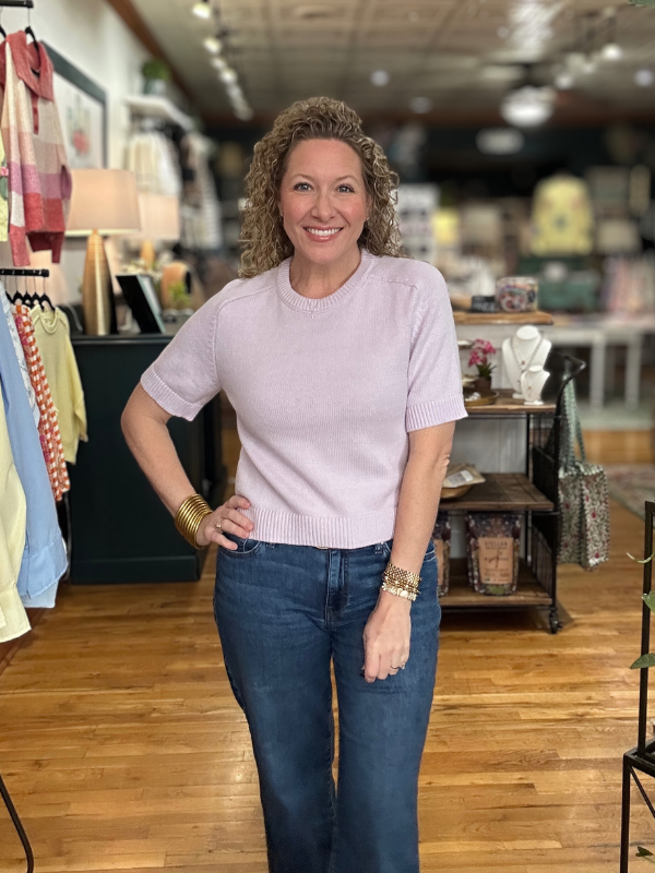 Woman in a light pink sweater and blue jeans standing in a store with wooden floors and clothing racks.