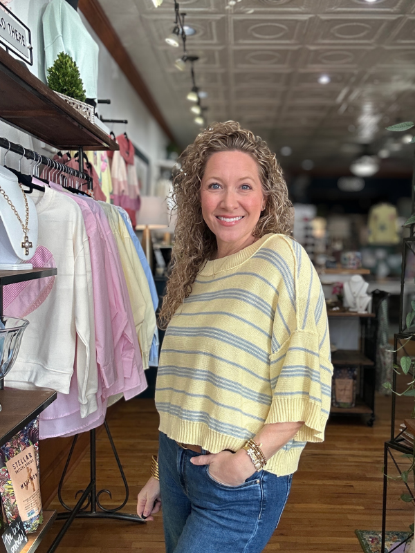 Woman in a yellow striped sweater standing in a store with clothing racks and displays.
