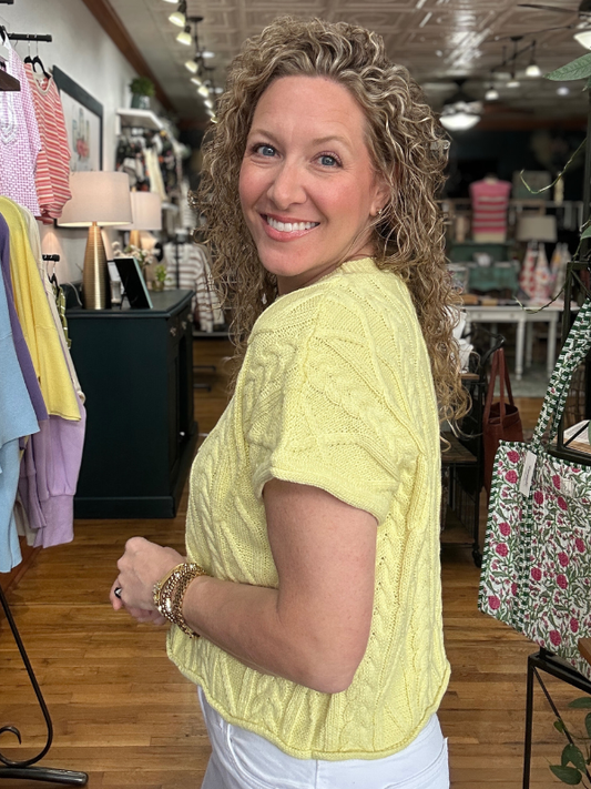 Woman in a yellow sweater standing in a store with clothes and bags around her.