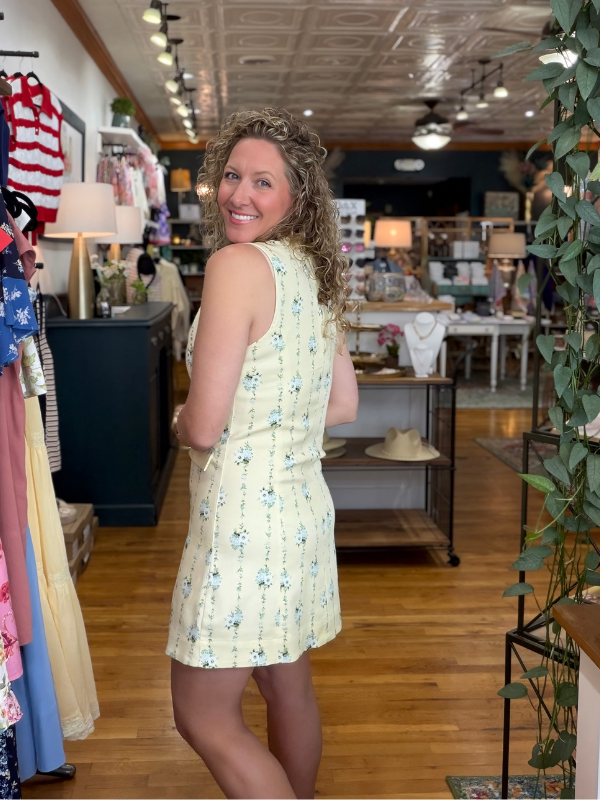 Woman in a yellow floral dress standing in a store with clothing racks and products in the background.