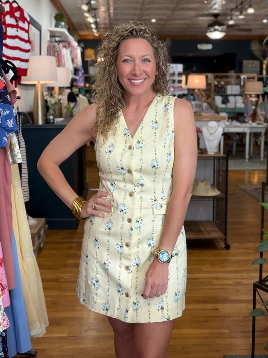 Woman in a yellow floral dress standing in a store with clothing racks and products in the background.