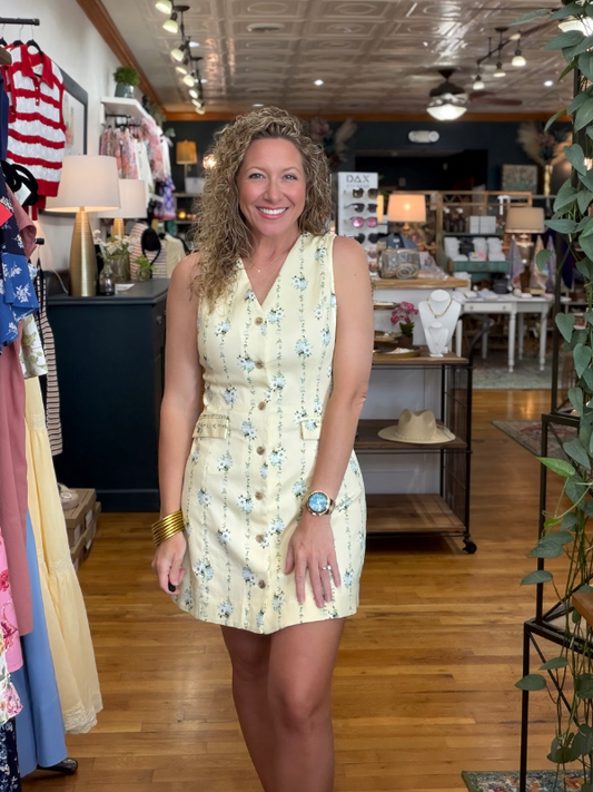 Woman in a yellow floral dress standing in a store with clothing racks and products in the background.