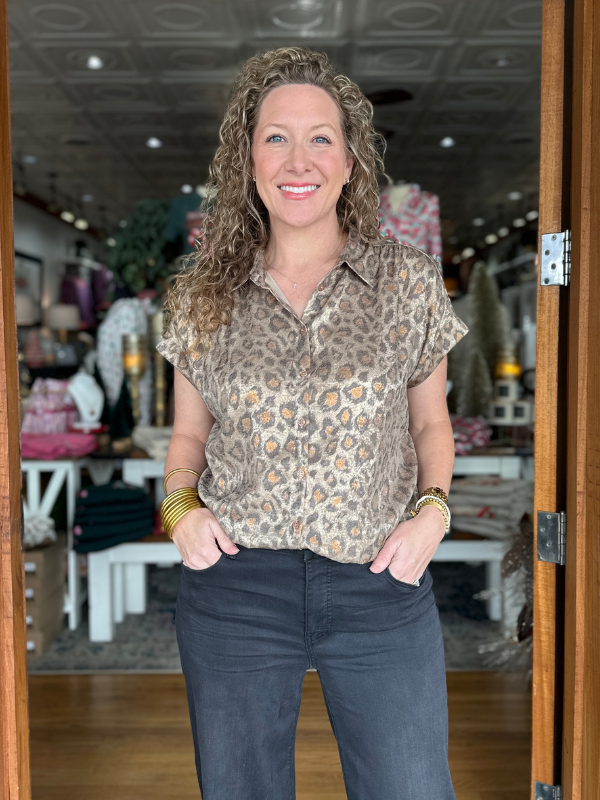 Woman standing in a store wearing a patterned blouse and jeans, with a blurred interior background.