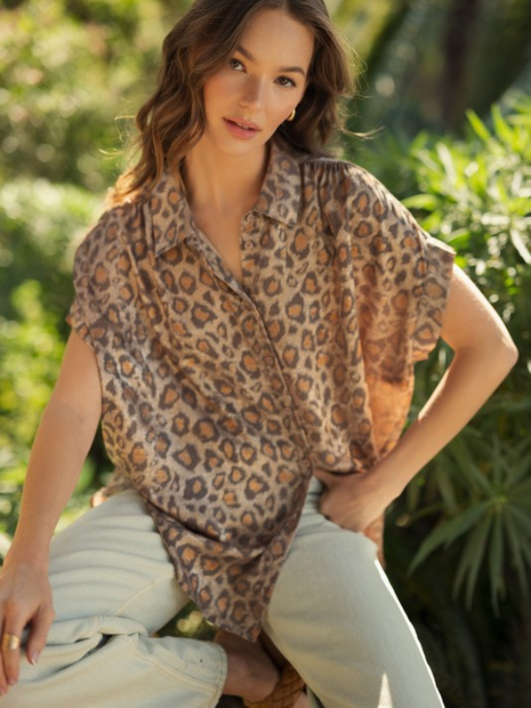 Woman wearing a leopard print top and light pants sitting outdoors with greenery in the background