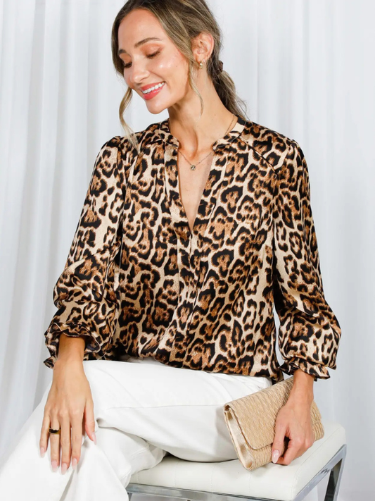 Woman wearing a leopard print blouse and white pants, sitting on a chair with a white curtain background.
