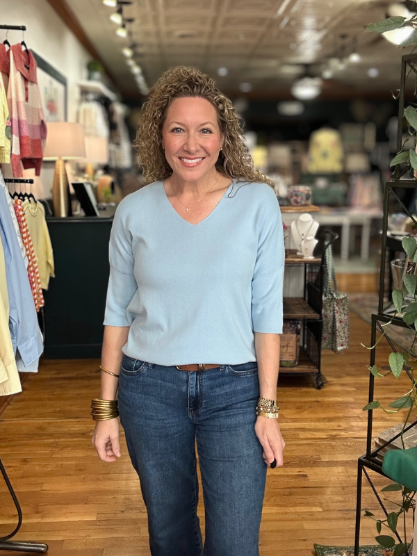 Woman in a light blue sweater and jeans standing in a store with wooden floors and clothing racks.