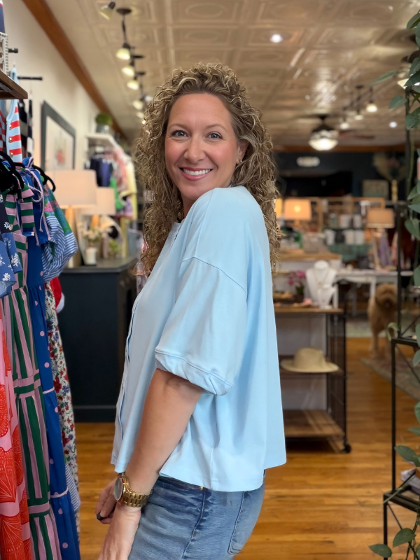 Woman in a light blue shirt standing in a store with clothing racks and decor.