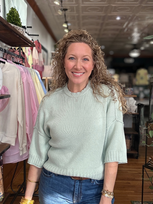 Woman wearing a light green sweater and blue jeans in a clothing store.