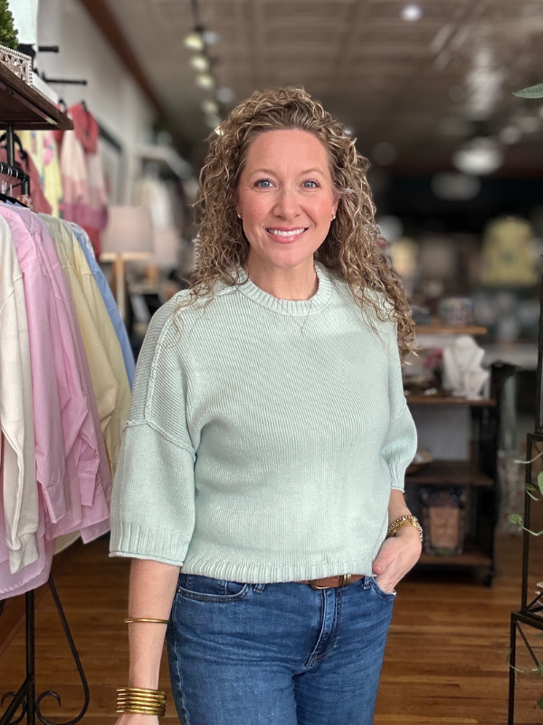 Woman wearing a light green sweater and blue jeans standing in a store with racks of clothes in the background.
