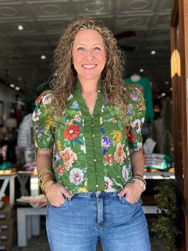 Woman wearing a green floral shirt and blue jeans standing indoors.
