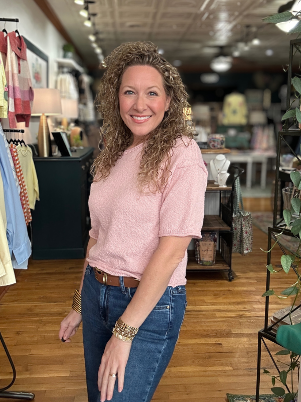 Woman in a pink shirt and blue jeans standing in a store with wooden floors and clothing racks.