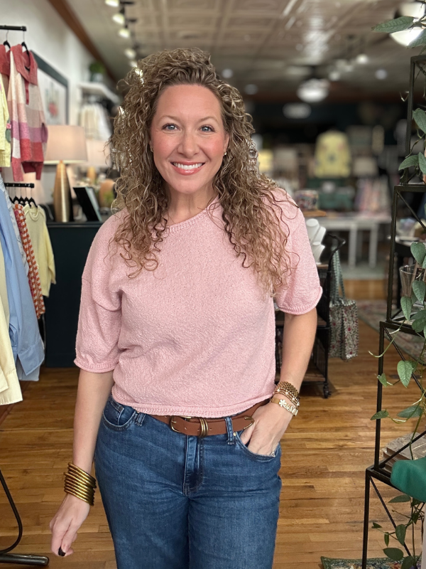 Woman in a pink shirt and blue jeans standing in a store with clothing racks in the background.