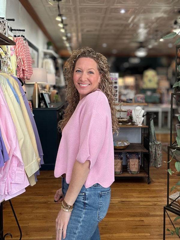 Woman in a pink top and blue jeans standing in a store with clothing racks and products in the background.