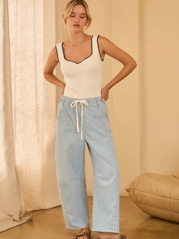 Woman wearing a white tank top and blue striped pants standing against a beige curtain.