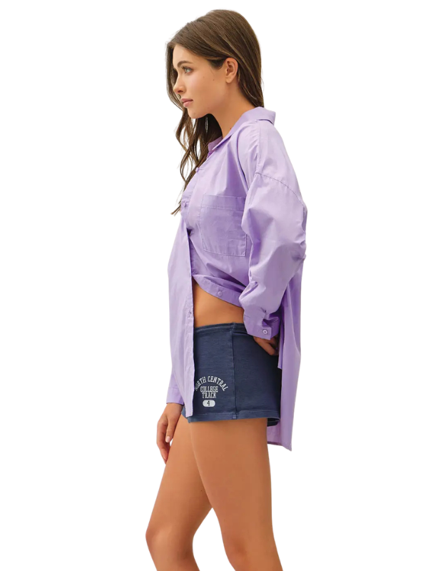 A woman shown in side profile wearing an oversized light purple/lavender button-up shirt and dark navy shorts with white collegiate-style text on the pocket, against a plain white studio background.