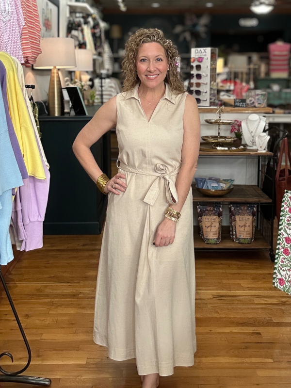 Woman in a beige dress standing in a store with clothing racks and products in the background.