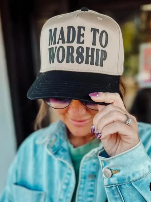 A woman wearing a beige and black trucker hat with the text 'MADE TO WORSHIP' in black lettering, paired with a light blue denim jacket and sunglasses.