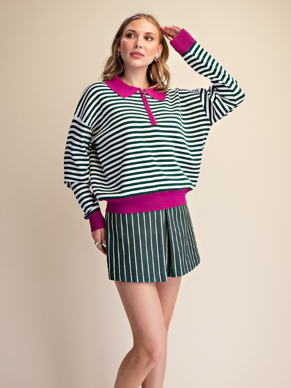 A woman modeling a matching two-piece outfit featuring a long-sleeve knit sweater with black and green horizontal stripes, a bright pink collar and hem, and matching green and white vertically striped shorts, set against a solid beige studio background.