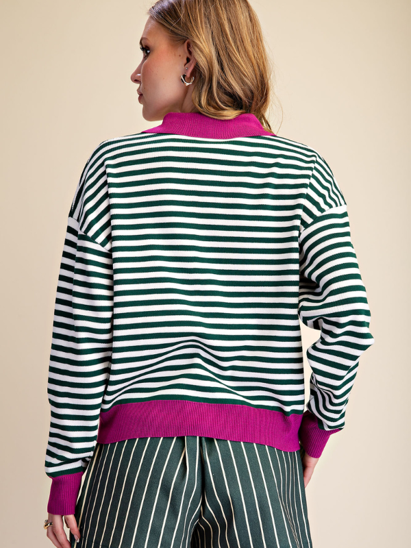 A woman shown from the back wearing a green and white striped knit sweater with bright magenta pink ribbed trim at the collar, hem, and cuffs, paired with dark green and white vertically striped pants, against a solid beige background.