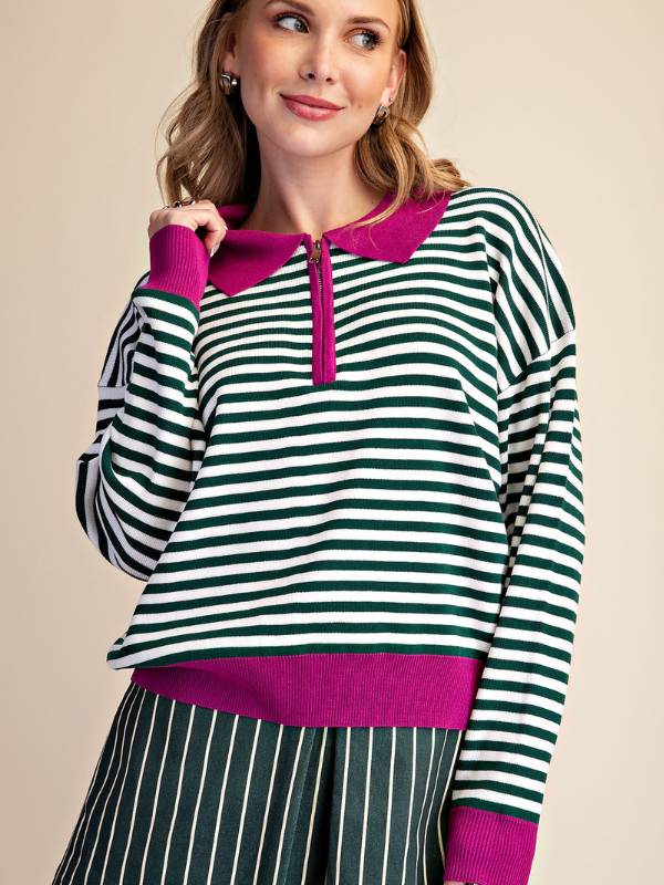 A woman wearing a green and white horizontally striped long-sleeve knit top with a bright magenta collar, zipper placket, hem, and cuffs, paired with matching green and white vertically striped bottoms, against a solid beige background.
