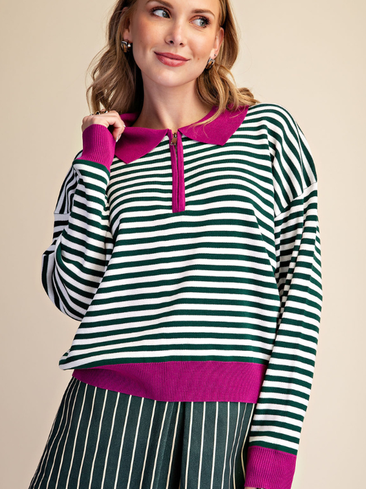 A woman wearing a green and white horizontally striped long-sleeve knit top with a bright magenta collar, zipper placket, hem, and cuffs, paired with matching green and white vertically striped bottoms, against a solid beige background.