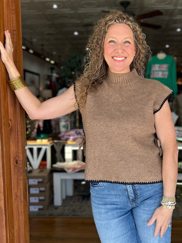 Woman wearing a brown sleeveless sweater and blue jeans standing indoors.