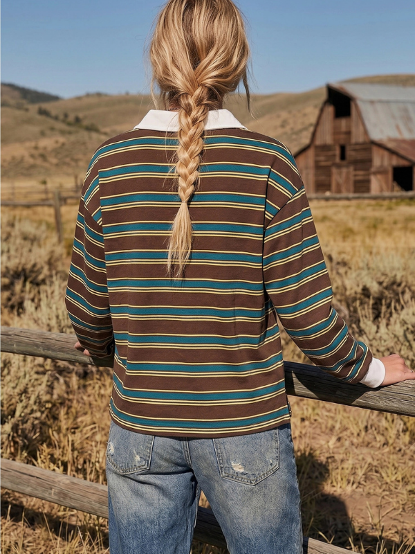 A woman viewed from behind wearing a brown, teal, and beige striped long-sleeve shirt with a white collar, standing in a rural outdoor setting with a wooden fence and a barn in the background.