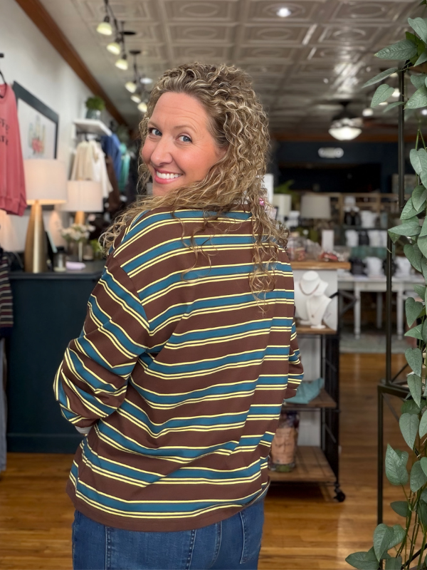 A woman with curly blonde hair is shown from the back wearing a brown, teal, and yellow striped long-sleeve shirt inside a retail store.