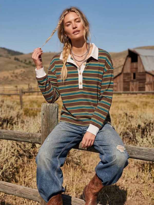 A woman sitting on a wooden fence in a rural outdoor setting, wearing a long-sleeve collared knit top with horizontal stripes in brown, green, and cream, paired with distressed blue jeans and brown boots.