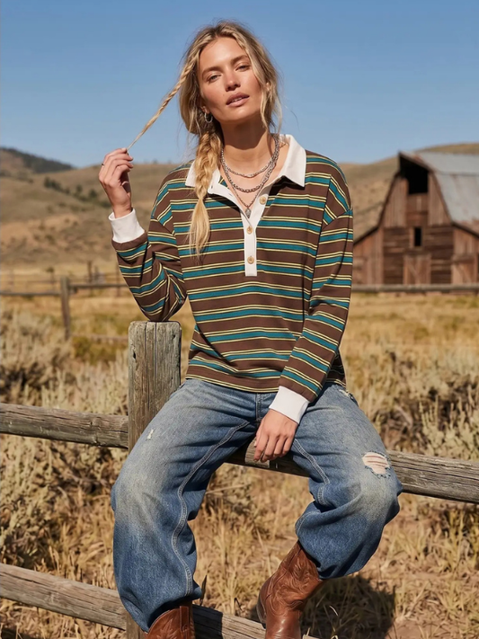 A woman sitting on a wooden fence in a rural outdoor setting, wearing a long-sleeve collared knit top with horizontal stripes in brown, green, and cream, paired with distressed blue jeans and brown boots.
