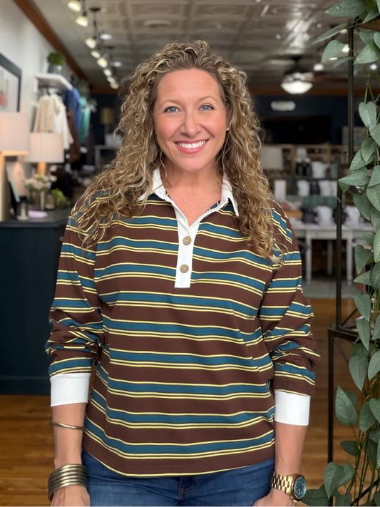 A smiling woman with curly blonde hair stands in a retail store wearing a brown, teal, and yellow striped long-sleeve polo shirt with a white collar and button placket, paired with blue jeans.