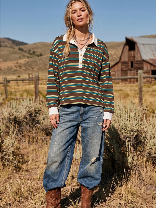 A woman stands outdoors in a rural landscape wearing a long-sleeve collared knit top with horizontal brown, teal, and cream stripes, paired with wide-leg blue jeans and brown boots.