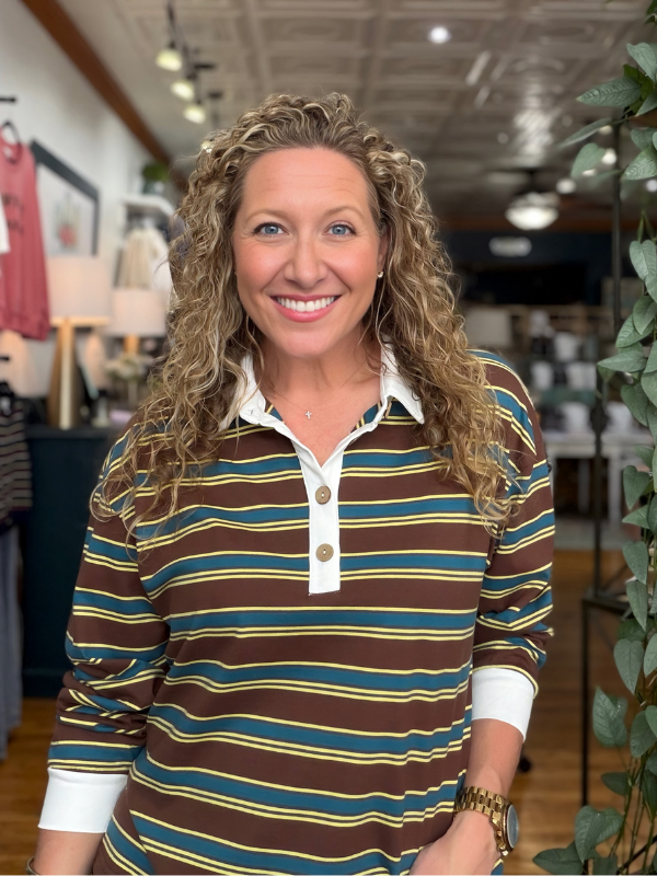 A smiling woman with long curly blonde hair wearing a brown, teal, and yellow striped long-sleeve polo shirt with a white collar, standing inside a retail store.