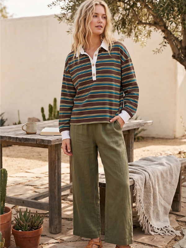A woman stands outdoors wearing a long-sleeve collared knit top with horizontal stripes in teal, brown, and cream, paired with olive green wide-leg pants. She is posed next to a rustic wooden table in a sunlit patio setting.