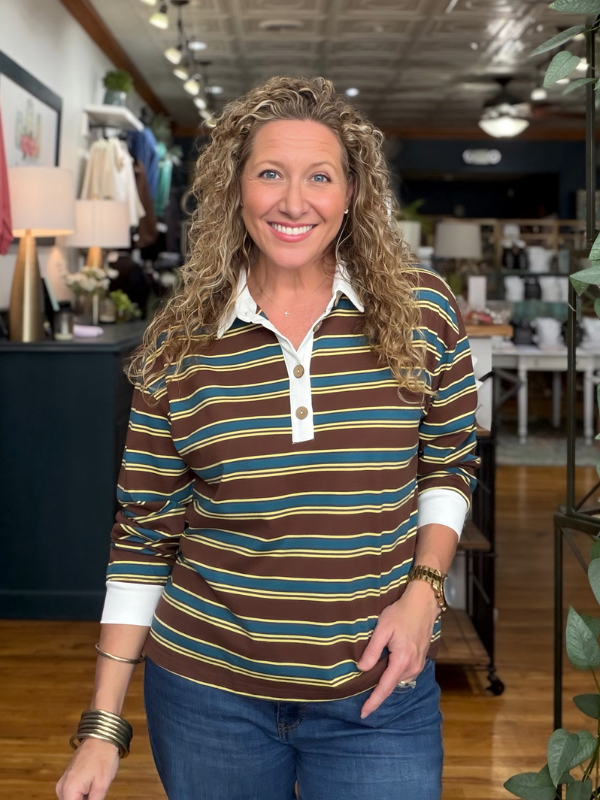 A smiling woman with curly blonde hair wearing a brown, teal, and yellow striped long-sleeve polo shirt with a white collar, standing in a retail store.