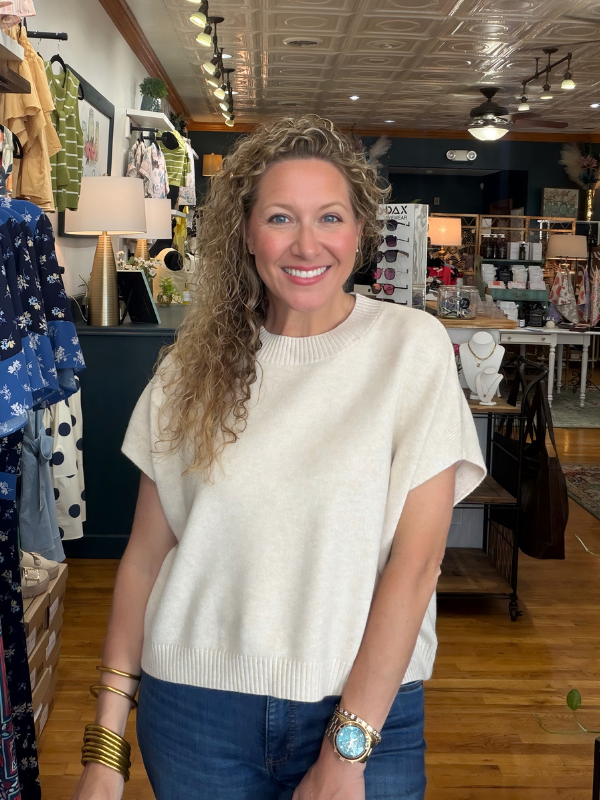 Woman in a store wearing a white sweater and blue jeans, smiling.
