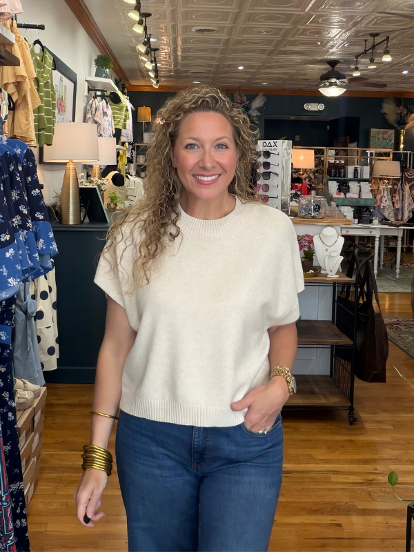 Woman in a store wearing a white sweater and blue jeans, smiling.